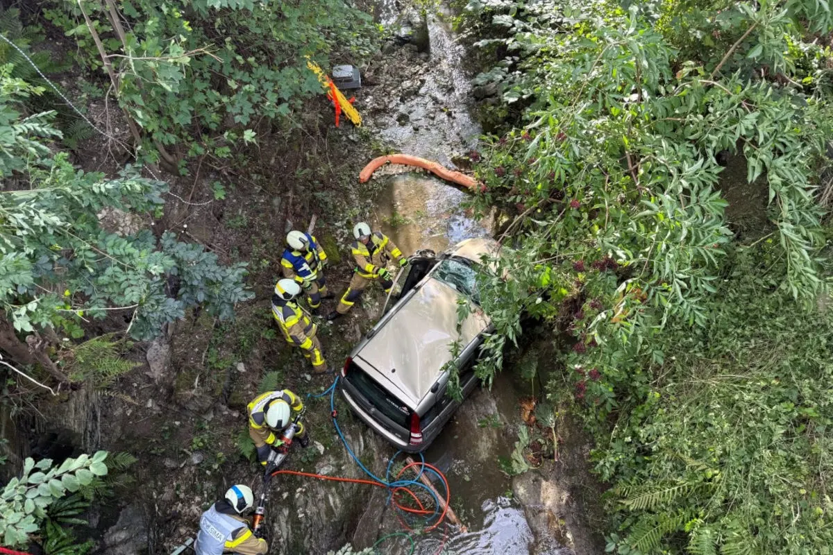 Ein Auto liegt in einem Bachbett, umgeben von Bäumen und Feuerwehrleuten in gelben Schutzanzügen. Diese sind dabei, das Fahrzeug zu sichern und bergen.