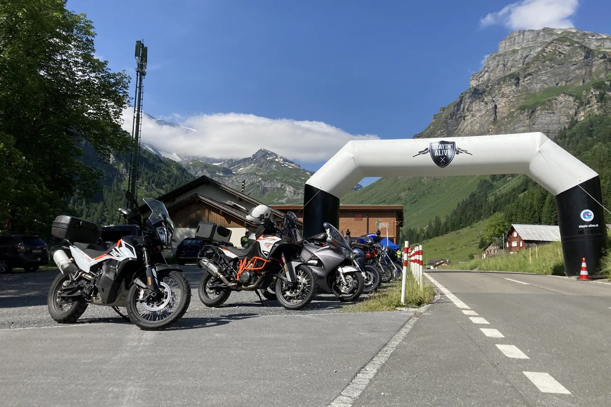 Vor einem weißen Bogen mit der Aufschrift ‚STAYIN' ALIVE X‘ stehen mehrere Motorräder geparkt. Die Umgebung ist alpines Gelände mit grünen Hügeln und blauen Himmel.