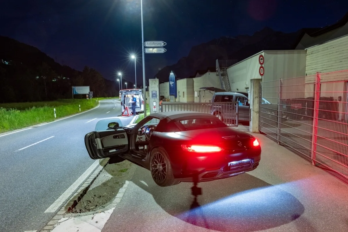 Ein schwarzer Mercedes steht mit offenen Fahrertür und leuchtenden Rücklichtern auf einer Landstraße neben einem Zaun. Im Hintergrund ist ein weiteres Fahrzeug zu sehen.