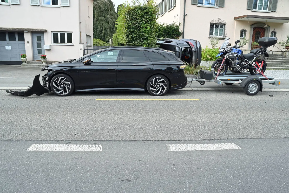 Ein schwarzes Auto mit einem beschädigten Vorderteil steht vor einem umgestürzten parkenden Auto. Ein Anhänger mit einem Motorrad ist angehängt.