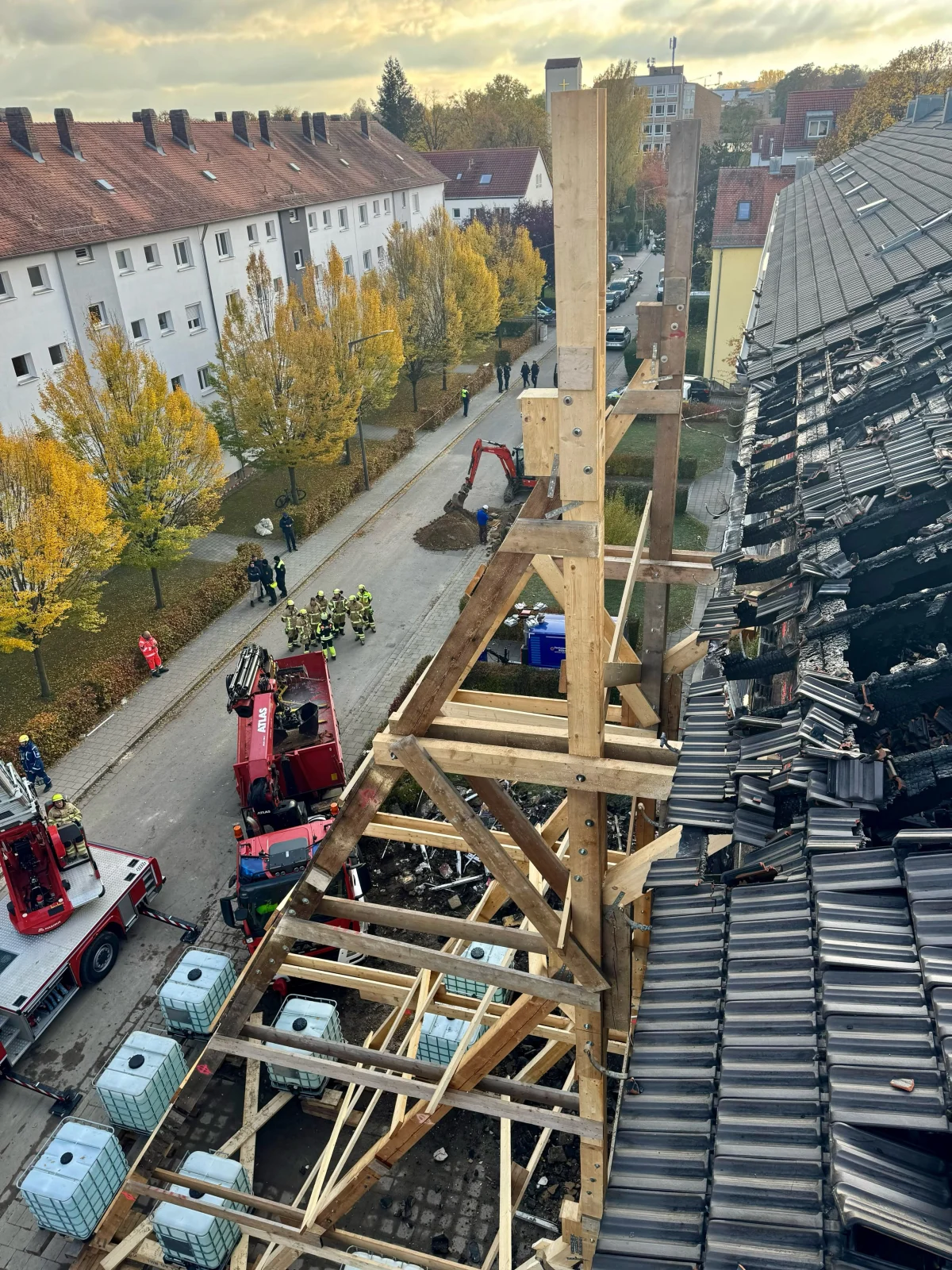Ein Baugerüst stützt ein teilweise zerstörtes Dach an einem Gebäude inmitten einer Wohngegend. Mehrere Einsatzfahrzeuge und Feuerwehrleute sind vor Ort.
