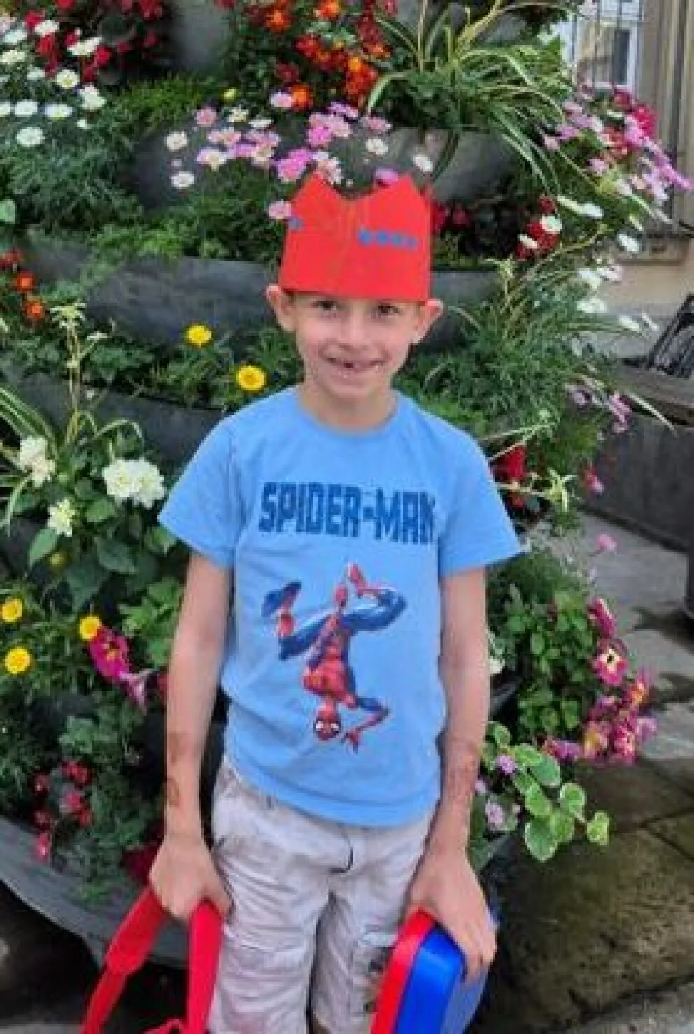Ein Junge trägt eine rote Papierkrone und ein blaues Spiderman-Shirt. Er steht vor einem bunten Blumenbeet.