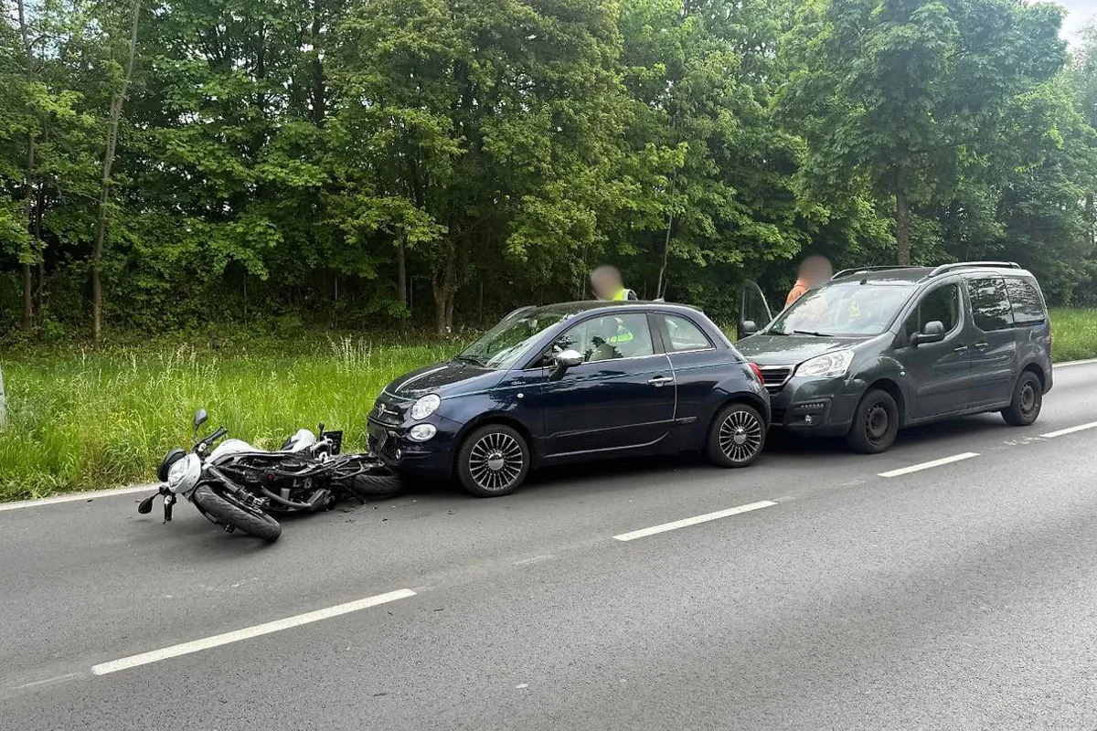 Auf der Straße liegt ein umgekipptes Motorrad vor zwei Autos. Die Szene befindet sich in einem bewaldeten Bereich.