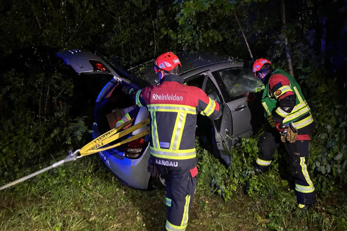 Zwei Feuerwehrleute mit Helmen und Reflektoranzügen bergen ein Auto, das in dichtem Gebüsch liegt. Auf ihren Jacken steht 'Rheinfelden' und 'AARGAU'.
