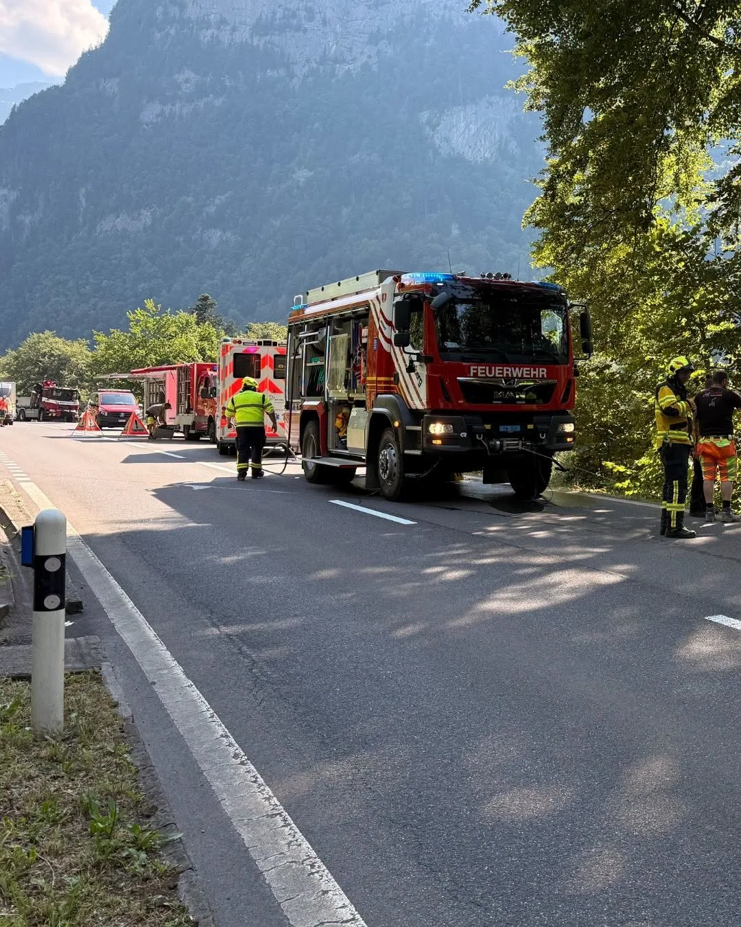 Ein rotes Feuerwehrfahrzeug steht auf einer Bergstraße, umgeben von mehreren Einsatzkräften und weiteren Einsatzfahrzeugen im Hintergrund.