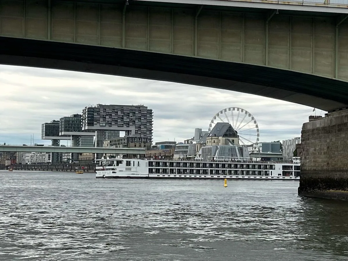 Ein Kreuzfahrtschiff fährt unter einer Brücke hindurch, im Hintergrund sind die Kranhäuser und ein Riesenrad in Köln zu sehen.