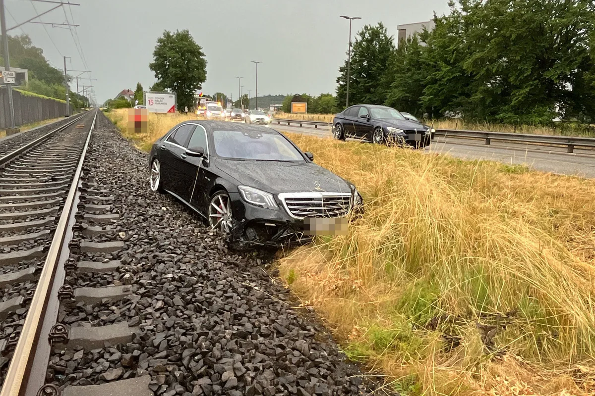 Ein schwarzer Mercedes steht schräg auf den Bahnschottern neben Bahngeleisen. Im Hintergrund ist eine Straße mit weiteren Fahrzeugen zu sehen.