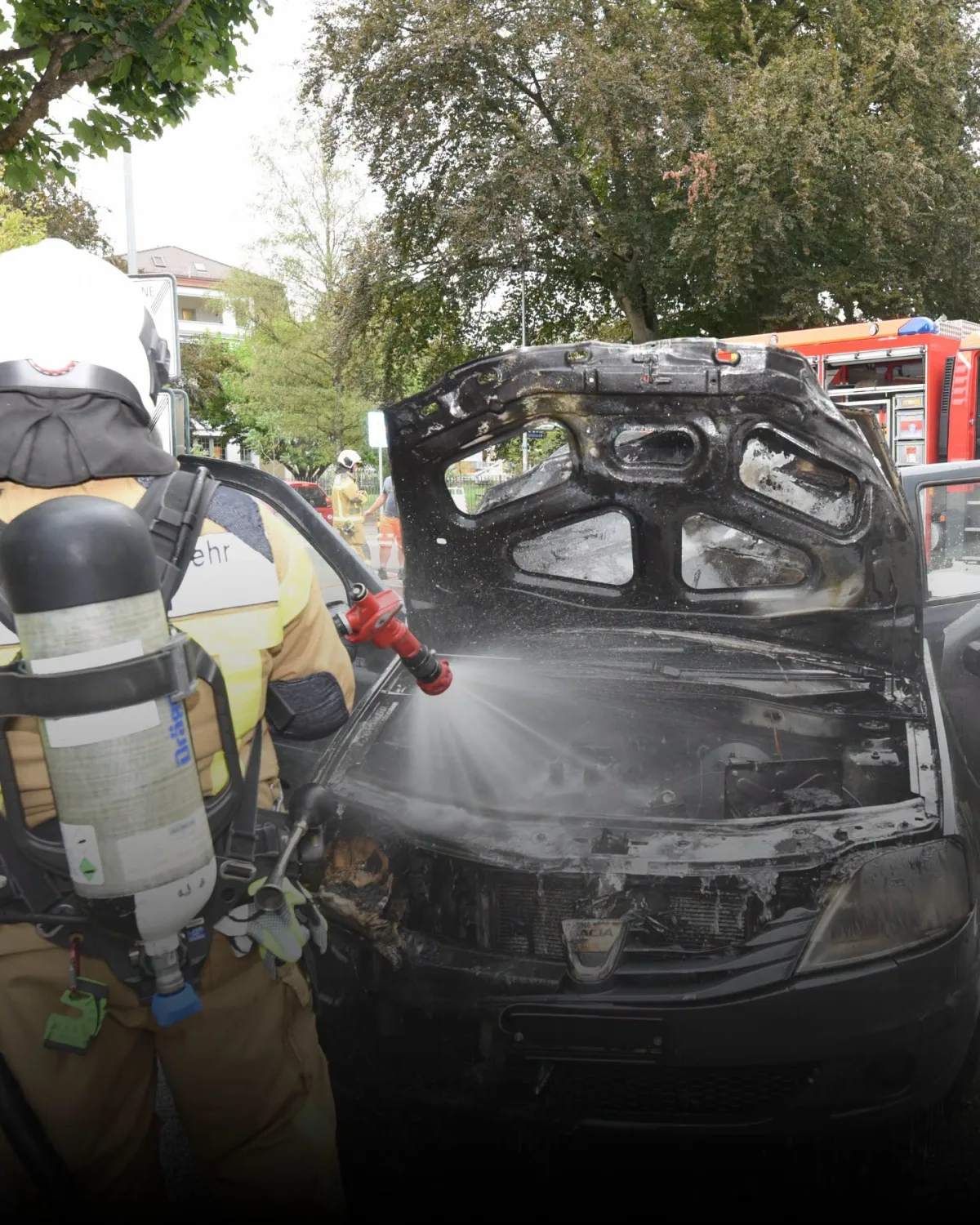 Ein Feuerwehrmann in Schutzkleidung löscht mit einem Wasserschlauch den Motorraum eines schwarzen Pkw. Der Fahrzeugbrand tritt in einem Wohngebiet auf, umgeben von Bäumen und Wohngebäuden.