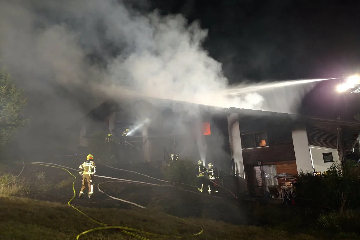 Mehrere Feuerwehrleute bekämpfen bei Nacht einen Brand in einem zweistöckigen Wohnhaus. Dicker Rauch steigt auf.