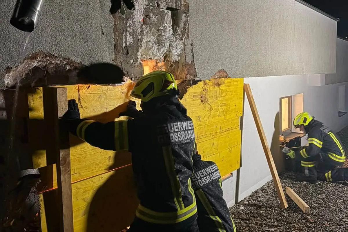 Zwei Feuerwehrmänner in Schutzkleidung arbeiten daran, eine beschädigte Wand mit Holzplanken zu sichern. Die Szenerie findet bei Nacht statt.
