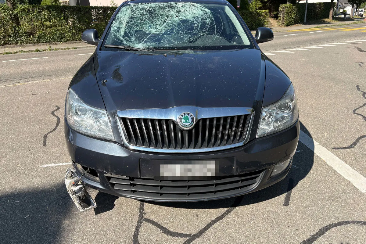 Ein dunkles Auto steht auf einer Straße. Die Windschutzscheibe ist stark beschädigt und der rechte Scheinwerfer hängt lose herunter.