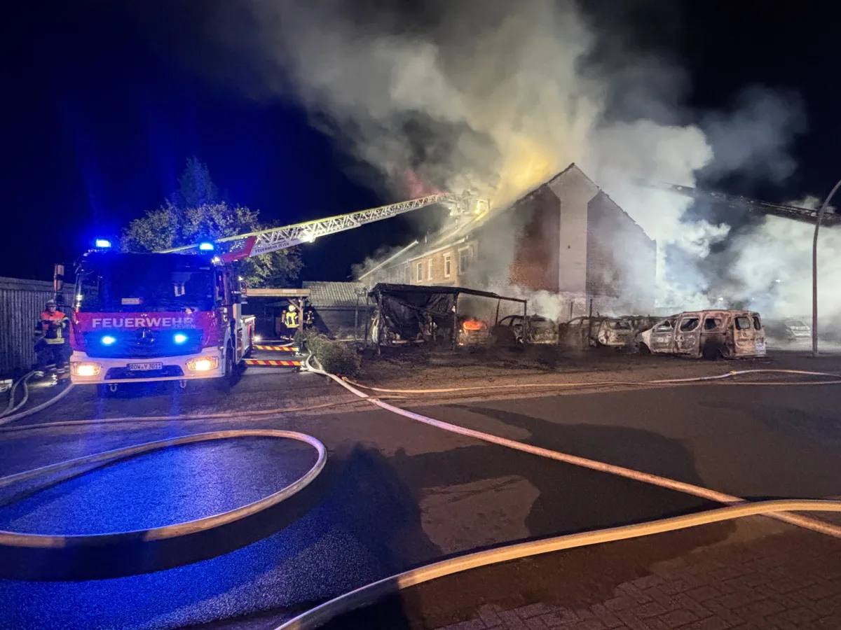 Ein großes Feuer brennt in einem Wohnhaus bei Nacht, während mehrere Feuerwehrleute mit einem Löschfahrzeug im Einsatz sind.