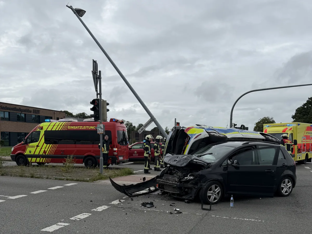 Ein schwarzes Auto mit frontalen Schäden steht neben einem umgekippten Rettungswagen an einer Kreuzung. Im Hintergrund sind Einsatzfahrzeuge sichtbar.