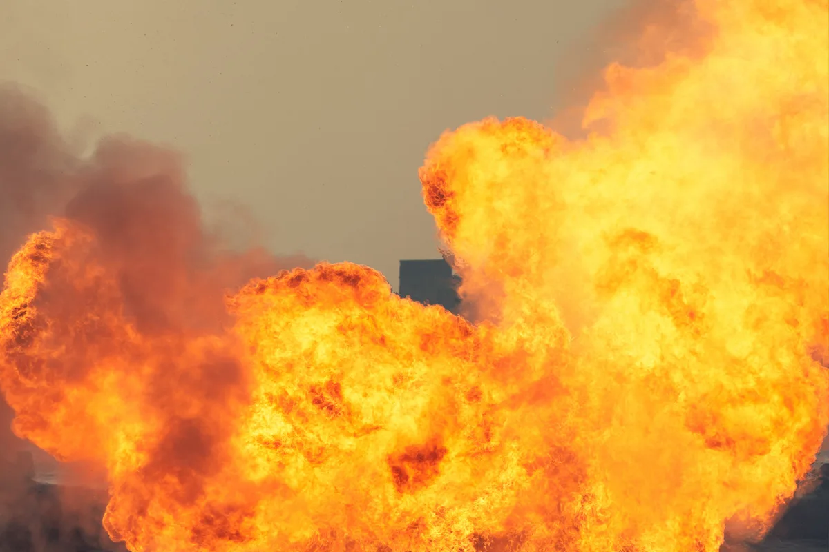 Das Bild zeigt große, helle Flammen und dichte Rauchwolken, die sich über den gesamten Bildausschnitt erstrecken. Der Hintergrund ist ein beiger Himmel, der die Konturen der Flammen und des Rauches hervorhebt.