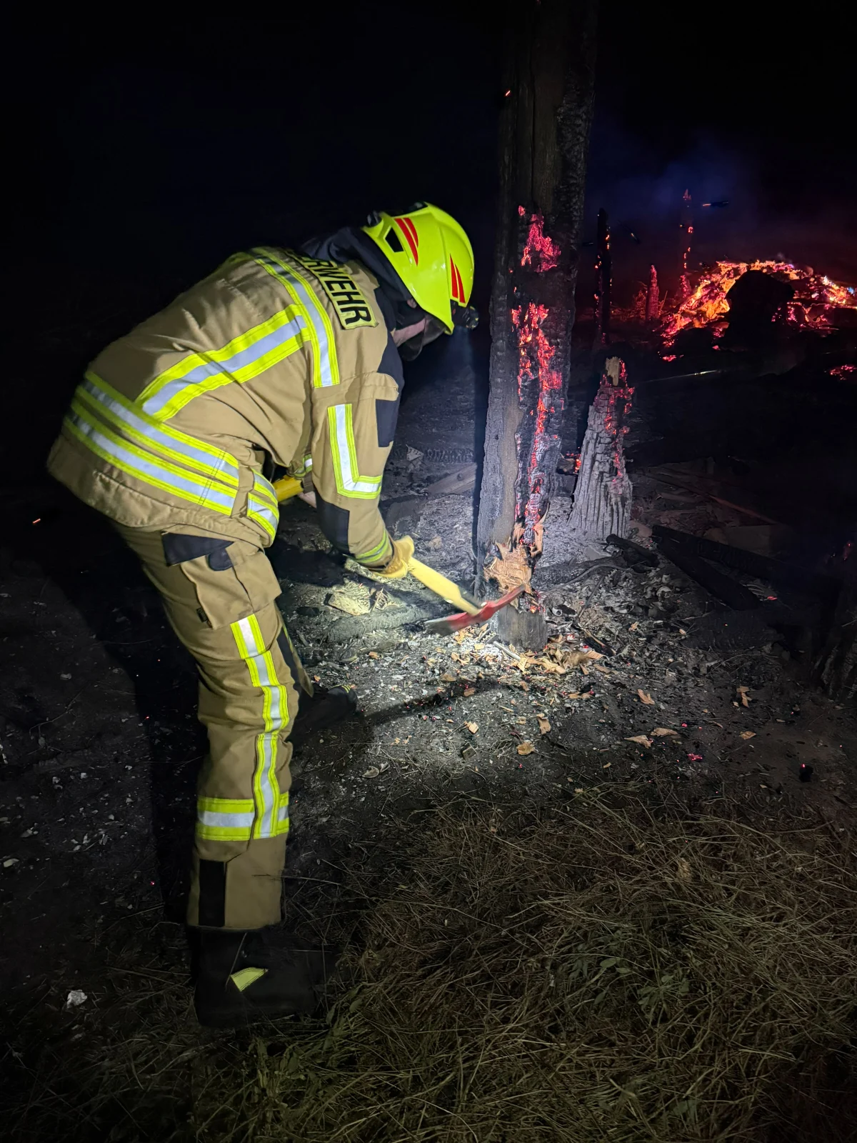 Ein Feuerwehrmann in Schutzkleidung mit Helm schlägt mit einer Axt auf glühendes Holz ein. Es ist Nacht, und das Umfeld ist dunkel.