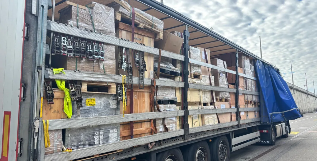 Ein Lastwagen steht mit geöffneter Plane am Straßenrand, beladen mit unterschiedlichen Holzkisten und Paletten, teilweise gesichert.