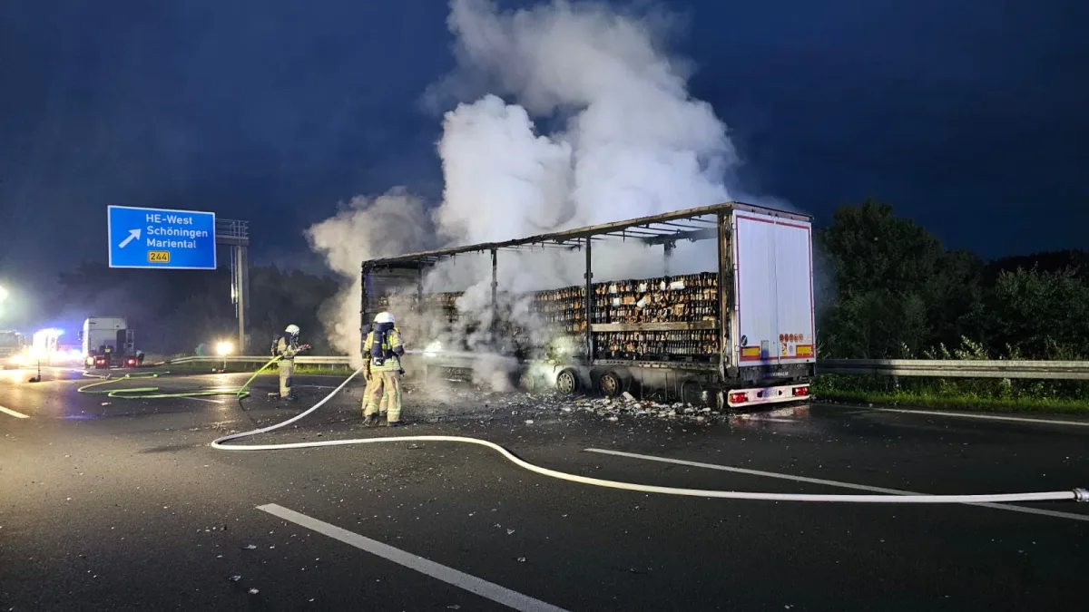 Auf einer Autobahn bei Nacht löschen Feuerwehrleute einen brennenden Lastwagen. Rauch steigt über der beschädigten Ladung auf.