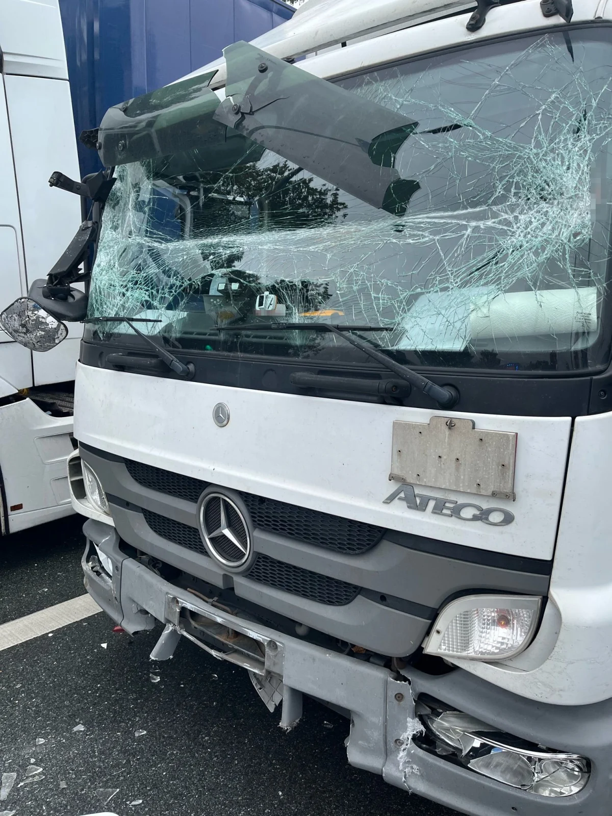 Ein weißer Mercedes-Lkw mit zerstörter Windschutzscheibe und schweren Frontschäden nach einem Unfall. Der Lkw steht auf einer Straße, daneben ist ein weiterer Lkw zu sehen.