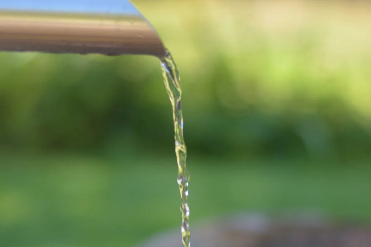 Ein kleiner Wasserstrahl fließt aus einem metallischen Ausguss in eine darunter liegende Grube. Der Hintergrund ist unscharf und grün.