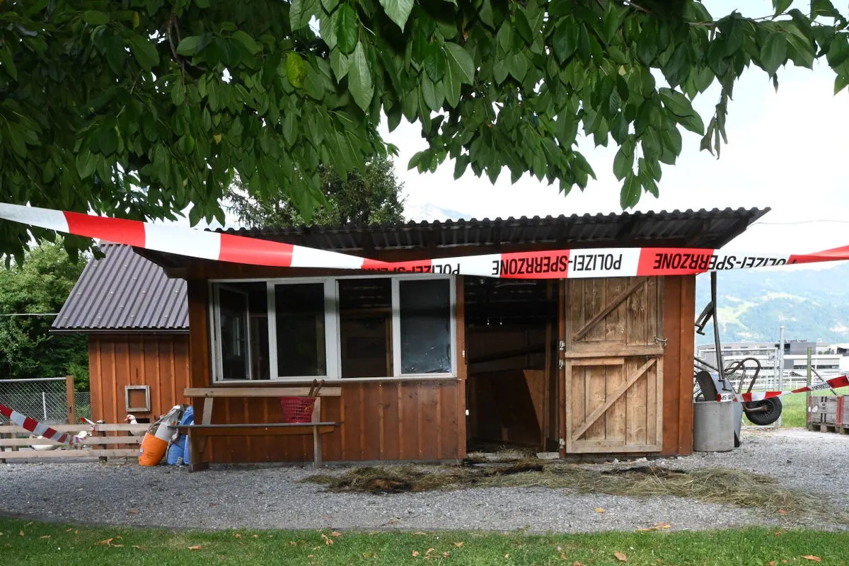 Ein ländlich gelegener Holzschuppen ist mit rot-weißem Polizeiband abgesperrt. Im Vordergrund sind Bäume und Wiesen zu sehen.