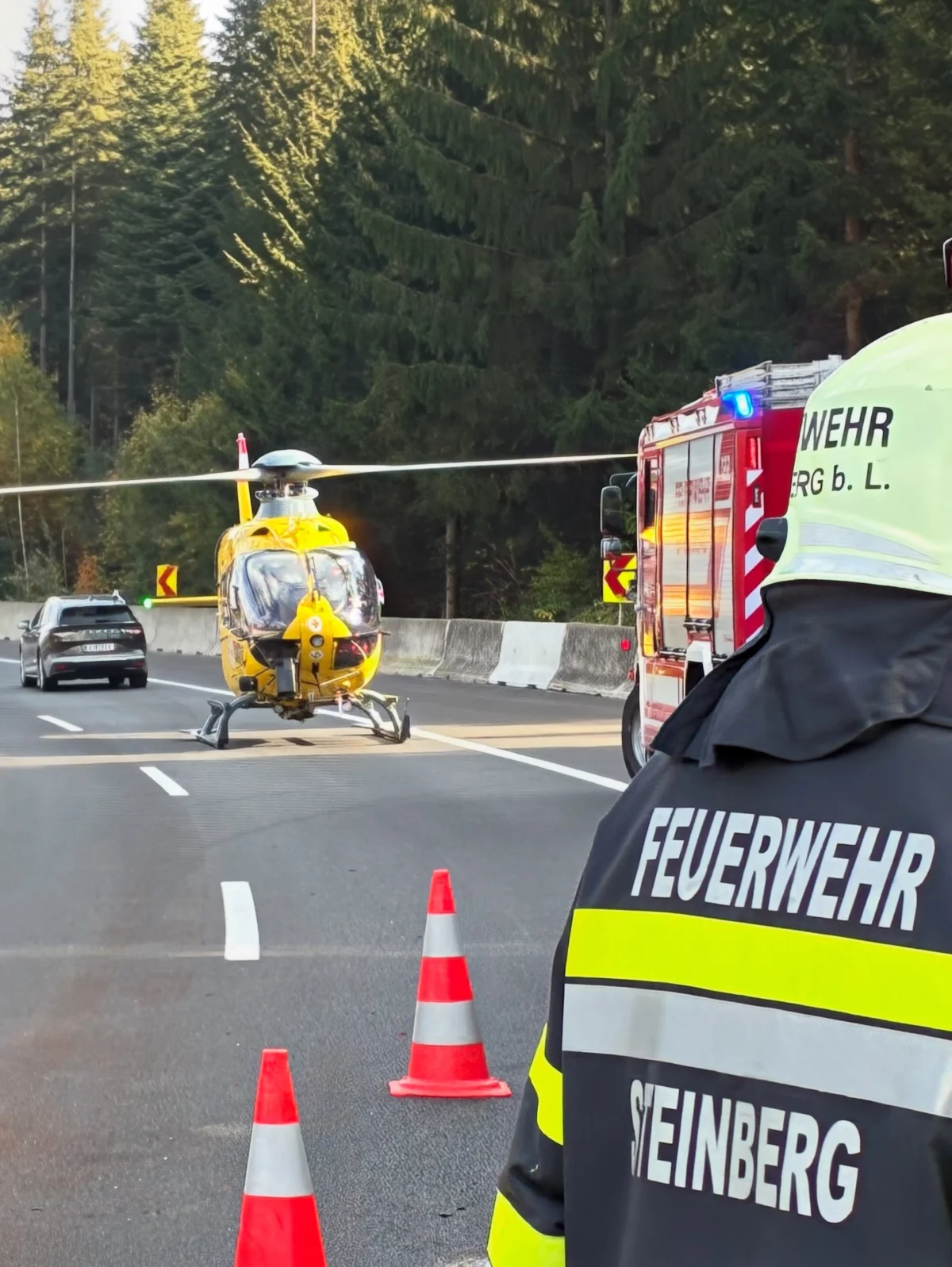 Ein gelber Rettungshubschrauber steht auf einer Autobahn. Im Vordergrund ist ein Feuerwehrmann mit der Aufschrift 'Feuerwehr Steinberg' auf der Jacke sichtbar.