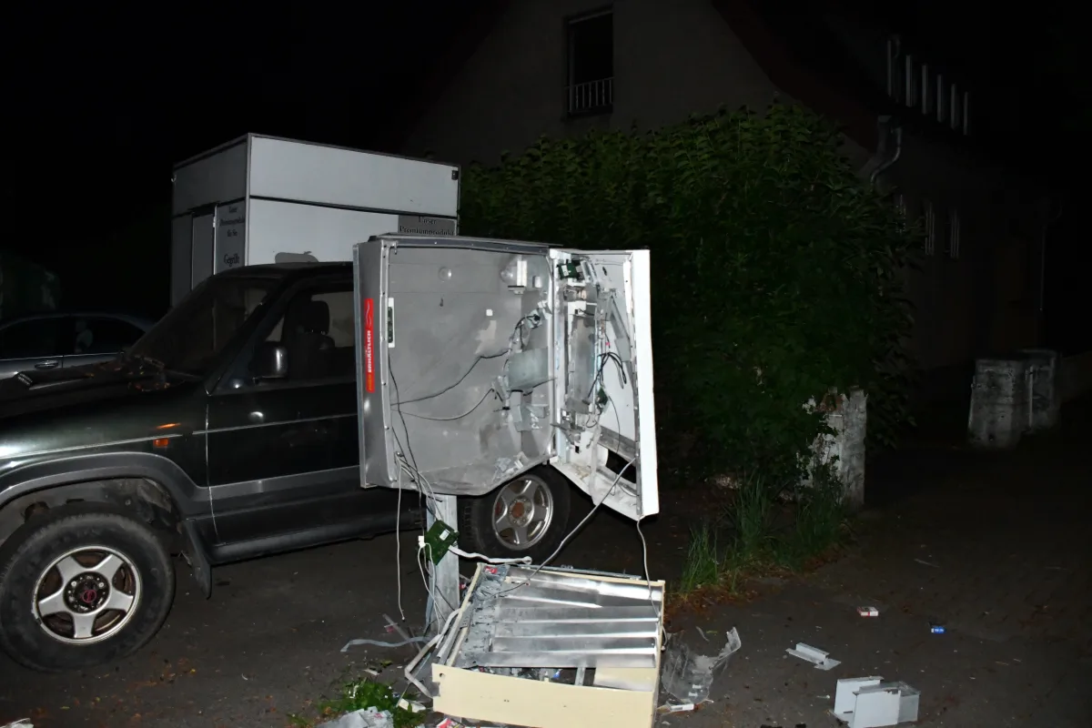 Ein Zigarettenautomat wurde zerstört und liegt auf dem Boden neben einem dunklen Fahrzeug. Gebäude und Vegetation sind im Hintergrund sichtbar.