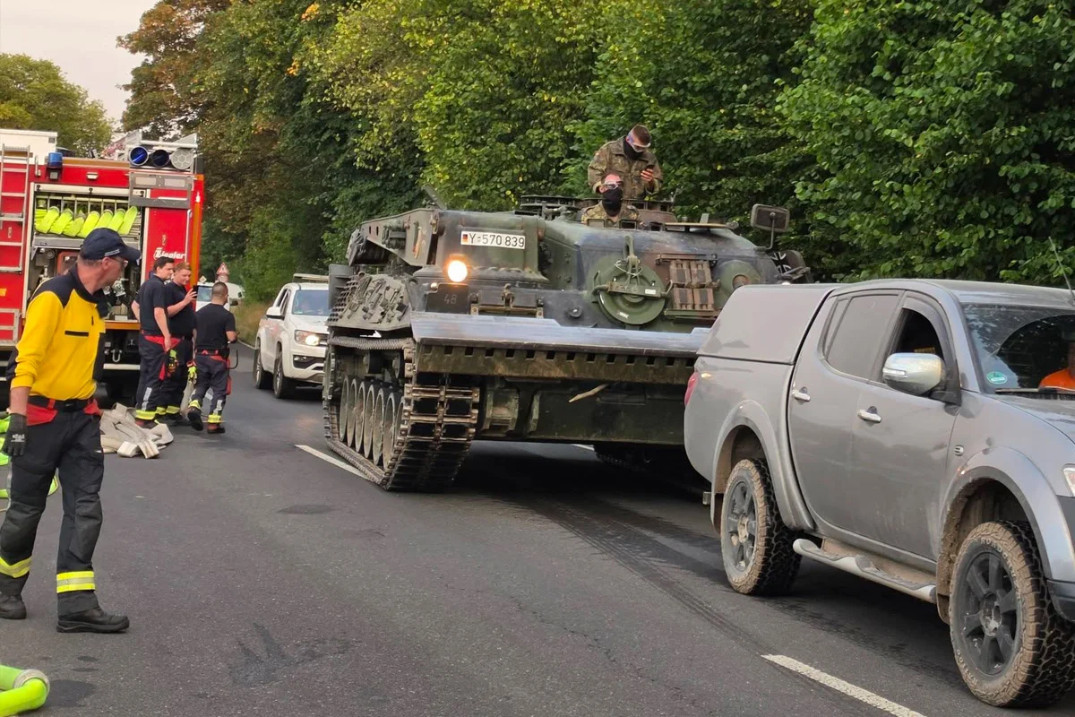 Ein Panzer steht auf einer Straße neben einem Feuerwehrauto mit Einsatzkräften in der Nähe und zivilen Fahrzeugen.