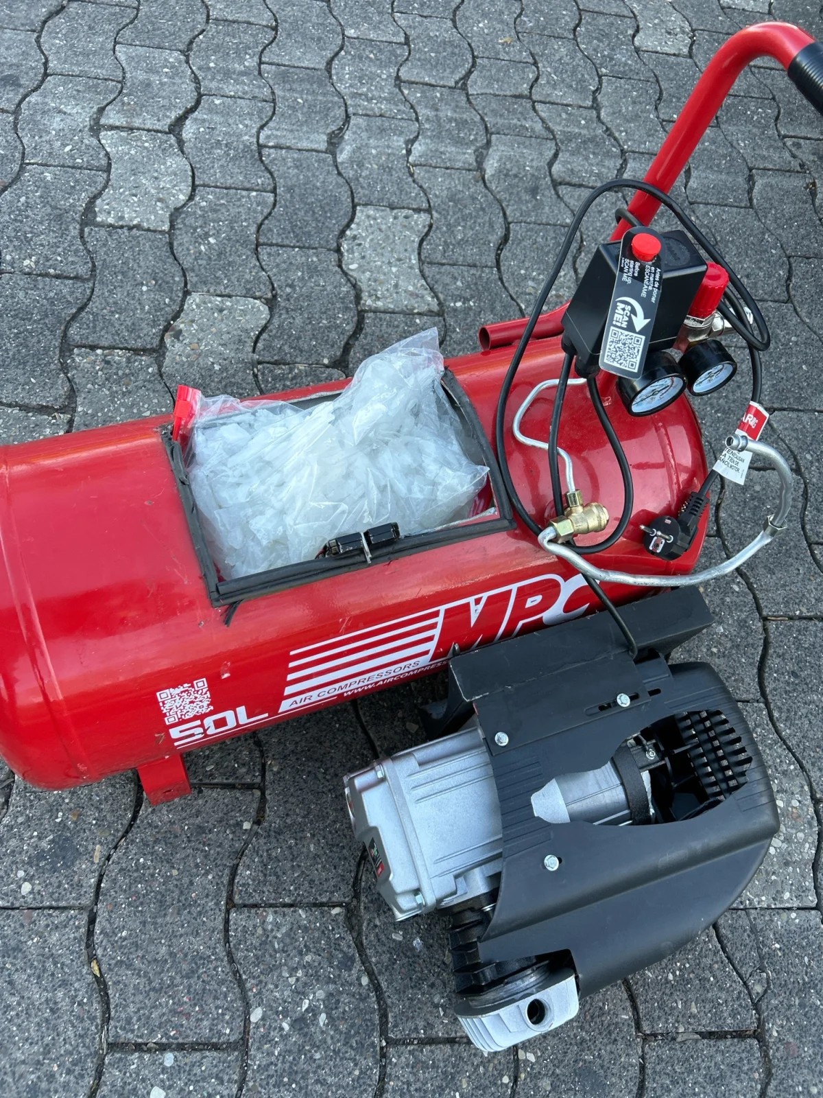 Ein roter Kompressor steht auf einem gepflasterten Boden. Das Fach des Kompressors ist geöffnet und enthält eine durchsichtige Plastiktüte.