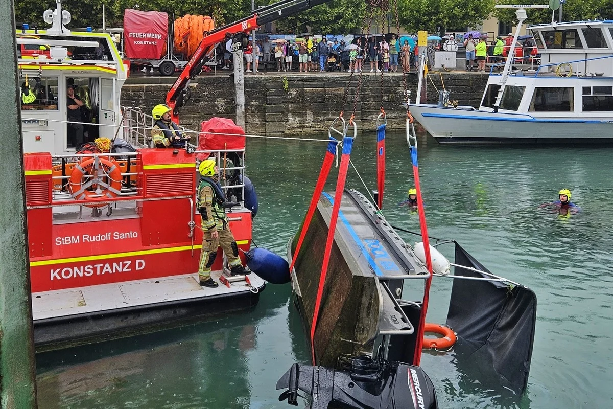 Feuerwehrleute heben ein Boot mit einem Kran aus dem Bodensee, während Zuschauer am Ufer zusehen. Zwei Feuerwehrtaucher sind im Wasser.