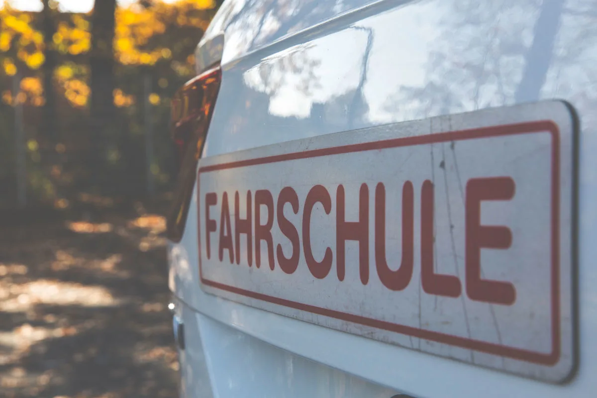 Ein Nahaufnahmebild zeigt ein 'Fahrschule'-Schild auf einem weißen Auto. Im Hintergrund sind herbstliche Bäume sichtbar.