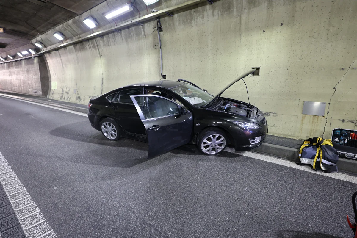 Ein schwarzes Auto steht in einem Tunnel mit offenen Türen und Kofferraumdeckel. Neben dem Auto liegen Gepäck und Werkzeugkoffer.