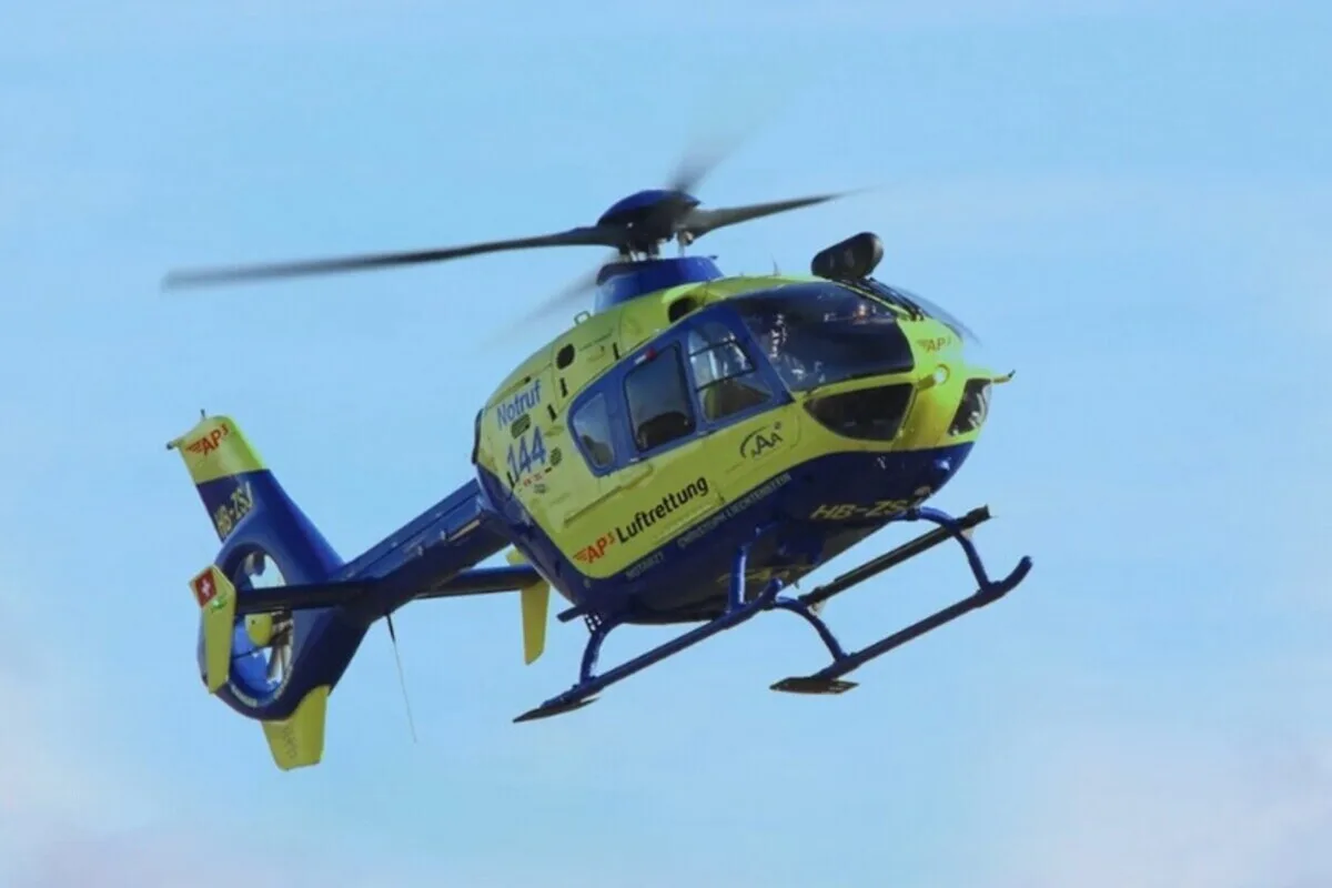 Ein gelb-blauer Rettungshubschrauber mit der Aufschrift 'Notruf 144' fliegt am lichtblauen Himmel. Der Hubschrauber trägt Logos der Luftrettungsdienste.
