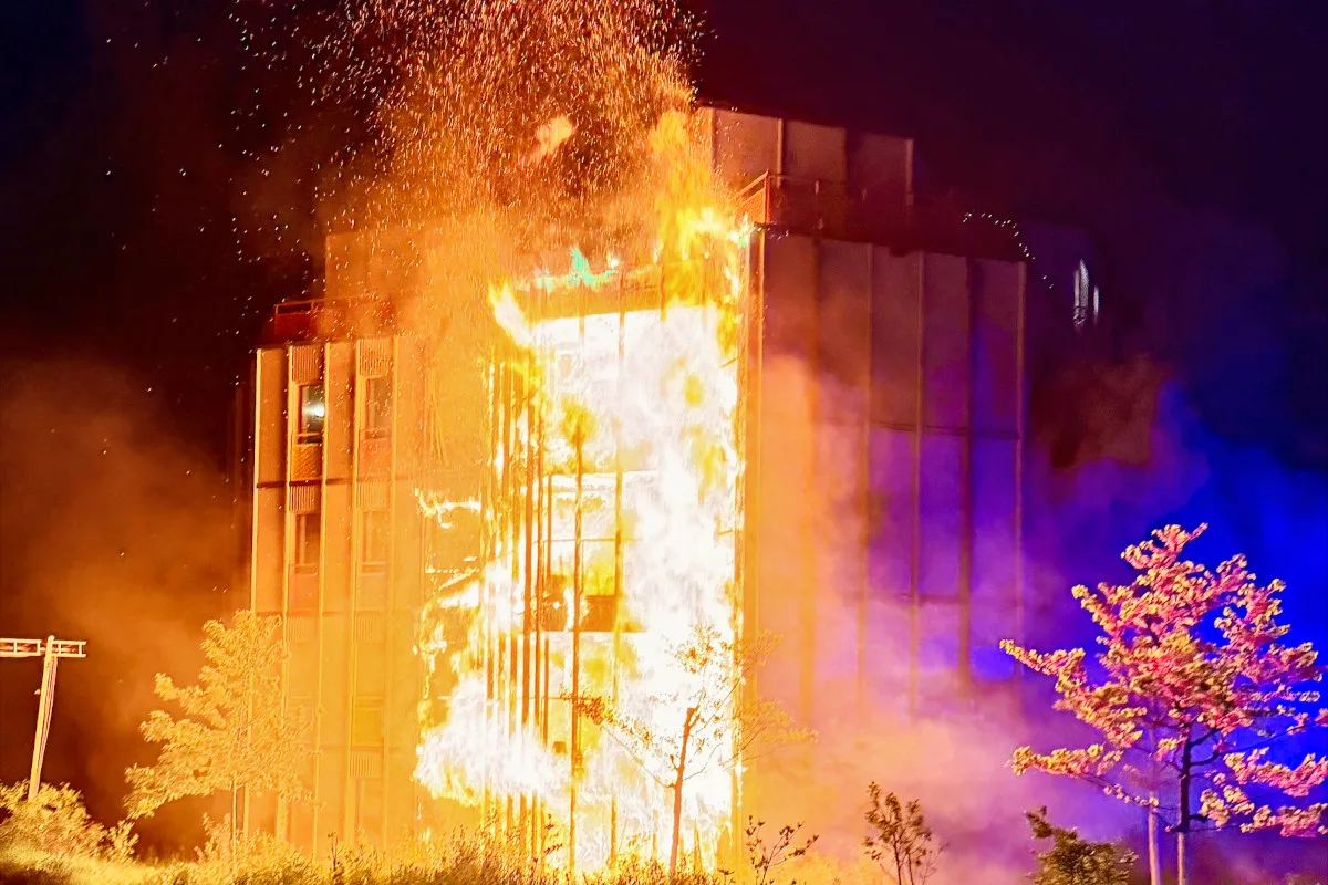 Ein großes, mehrstöckiges Gebäude brennt in der Nacht, Flammen und Funken sind deutlich sichtbar. Das Feuer erleuchtet die Umgebung, während Bäume im Vordergrund stehen.