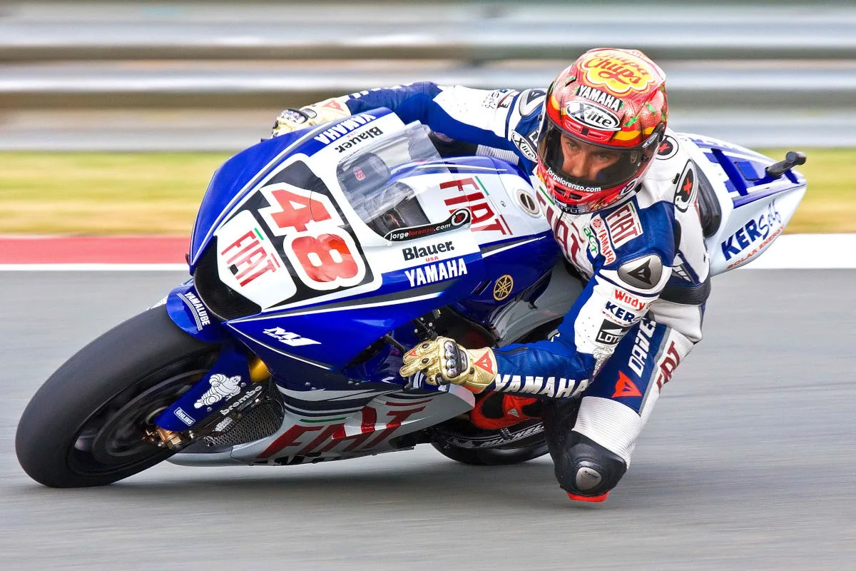 Ein Rennfahrer auf einer blauen Yamaha fährt in Schräglage eine Kurve auf einer Rennstrecke. Er ist in voller Schutzbekleidung und Helm.