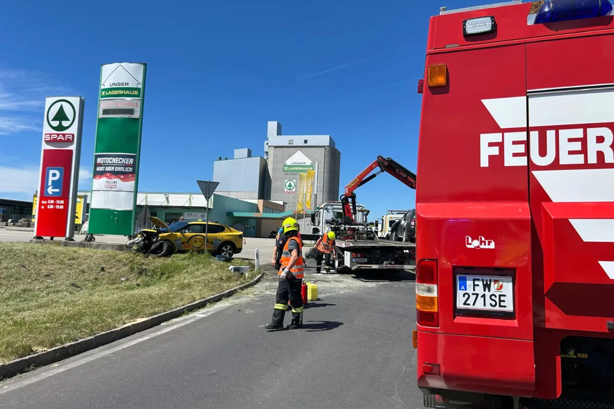 Ein Feuerwehrfahrzeug und Einsatzkräfte stehen an einer Unfallstelle, ein gelbes Auto ist beschädigt vor einem Supermarkt sichtbar.