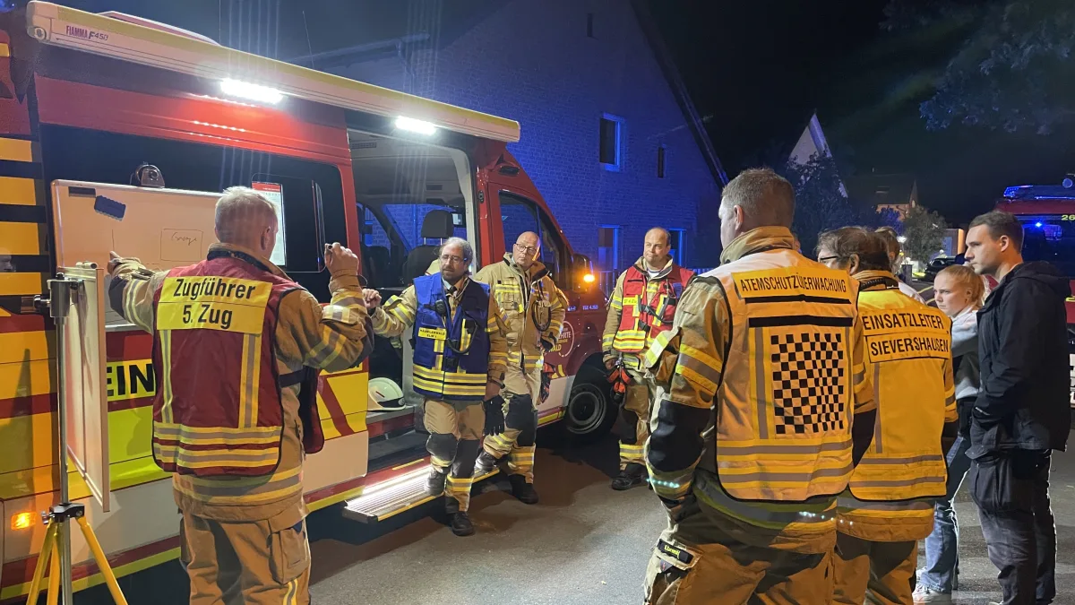 Mehrere Feuerwehrleute in Schutzkleidung stehen vor einem roten Einsatzfahrzeug und besprechen den Einsatz. Die Szene spielt bei Nacht in einem Wohngebiet.