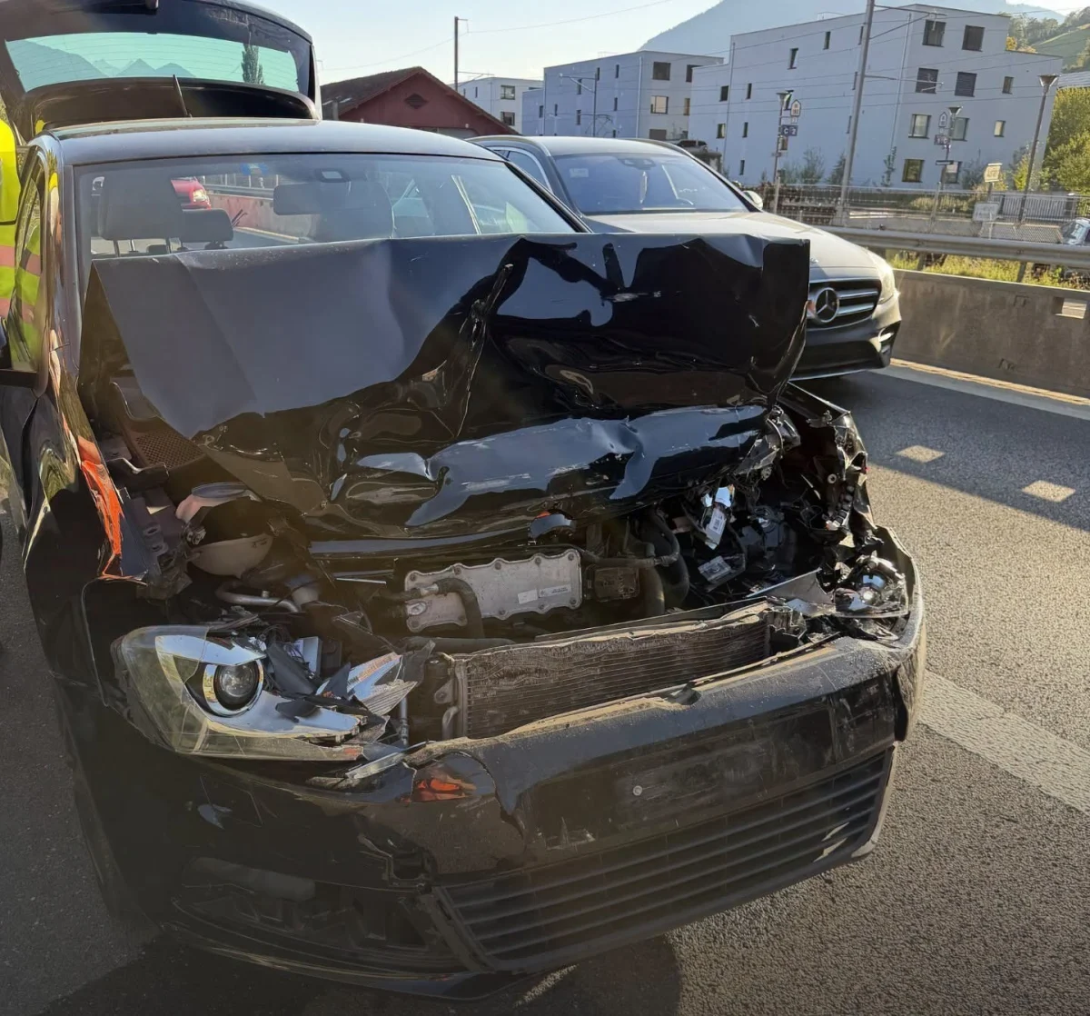 Ein schwarzes Auto mit massiven Schäden an der Front steht auf einer Straße. Neben dem Auto ist ein weiteres Fahrzeug sichtbar.