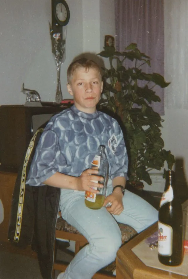 Eine junge Person sitzt in einem Wohnzimmer auf einem Stuhl und hält eine Flasche. Im Hintergrund sind Pflanzen und ein Regal sichtbar.