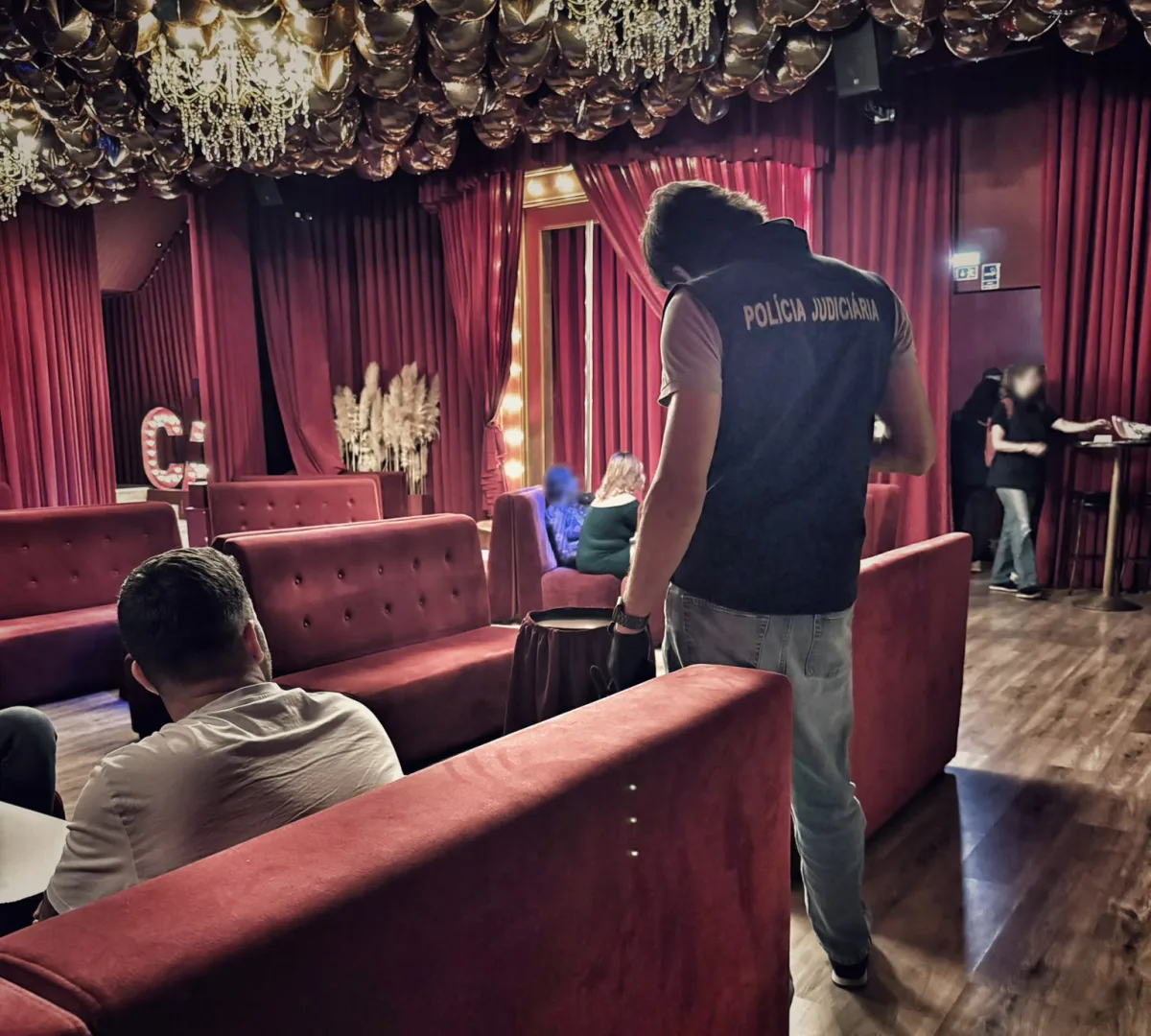Ein Ermittler steht in einem rot dekorierten Nachtclub mit einer Weste der Polícia Judiciária. Weitere Personen sind im Hintergrund sichtbar.