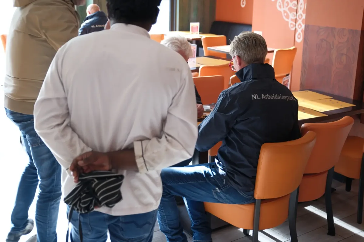Mehrere Personen sitzen und stehen in einem Restaurant. Eine Person trägt eine Jacke mit der Aufschrift 'NL Arbeidsinspectie'.