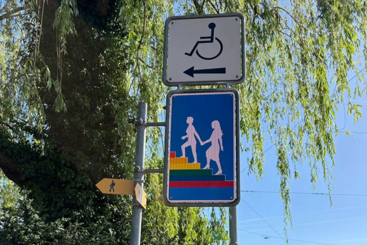 Zwei Schilder zeigen ein Rollstuhlsymbol und ein Symbol von Menschen, die eine bunte Treppe hinaufsteigen, umrahmt von grünem Laub.