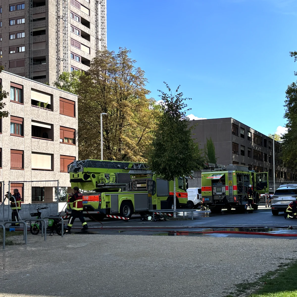 Mehrere Feuerwehrfahrzeuge sind vor einem hohen Wohngebäude im urbanen Gebiet im Einsatz. Feuerwehrleute sind aktiv sichtbar.