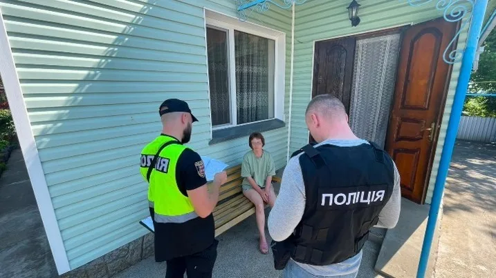 Zwei Polizisten, einer in gelber Weste mit der Aufschrift 'Polizei', sprechen mit einer Person, die auf einer Bank vor einem mintgrünen Haus sitzt.