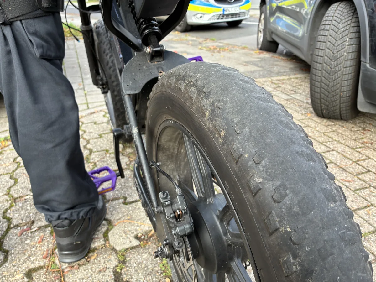 Im Vordergrund ist das Hinterrad eines robusten Fahrrads auf einem Gehweg zu sehen. Dahinter steht ein Polizeifahrzeug am Straßenrand.