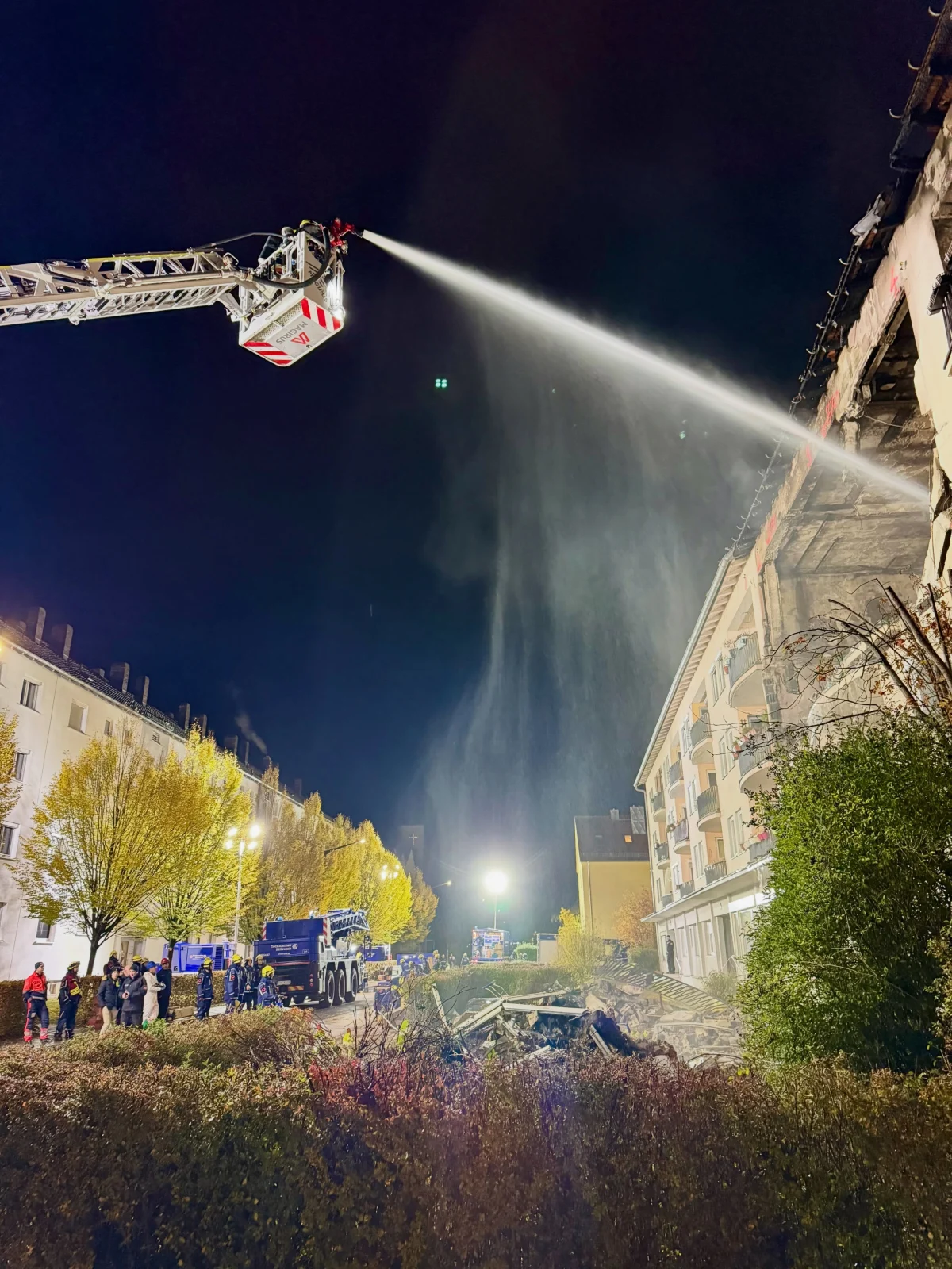 Ein Feuerwehrmann löscht von einer Hebebühne aus ein brennendes Gebäude bei Nacht. Menschen stehen neben Einsatzfahrzeugen.