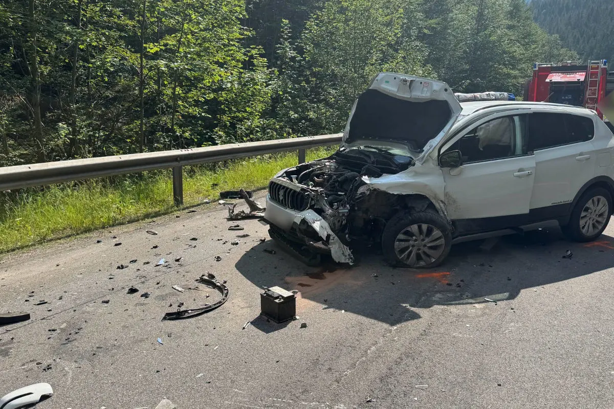 Ein weißes Auto mit schwerem Frontschaden steht auf einer Landstraße, umgeben von Wald. Fahrzeugteile liegen verstreut auf der Straße.