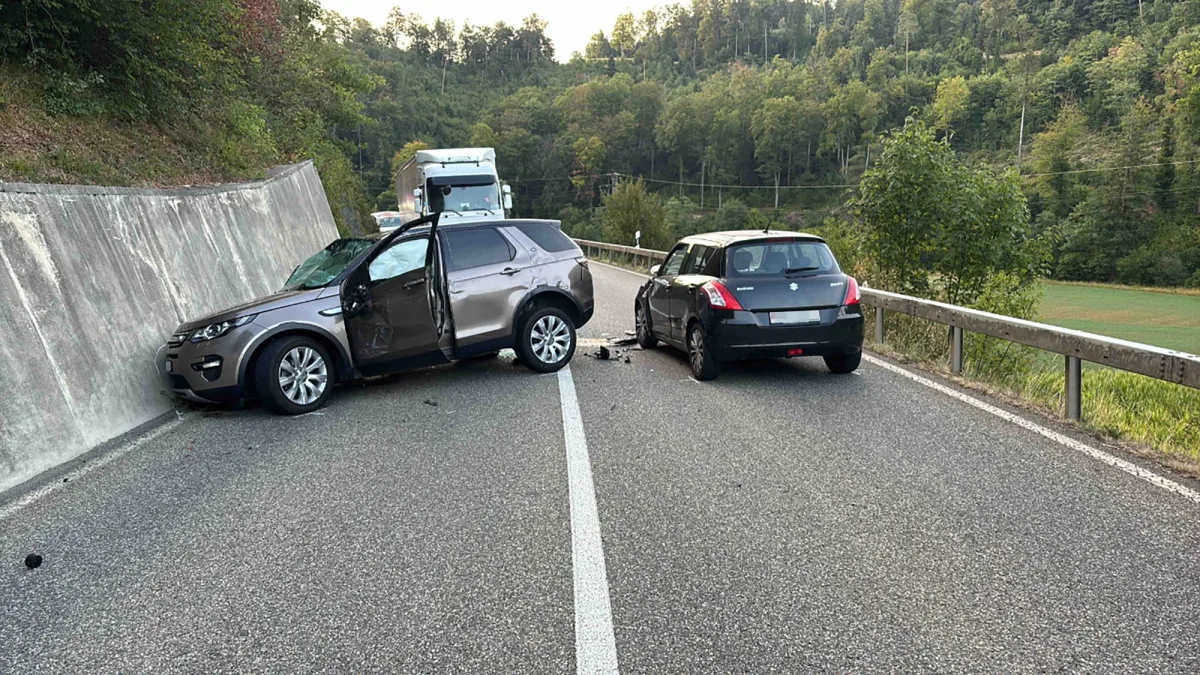 Ein graues SUV ist gegen eine Betonwand geprallt, während ein schwarzer Kleinwagen mit seinem Heck zur Kamera steht. Beide Fahrzeuge befinden sich auf einer Landstraße mit bewaldeter Umgebung.