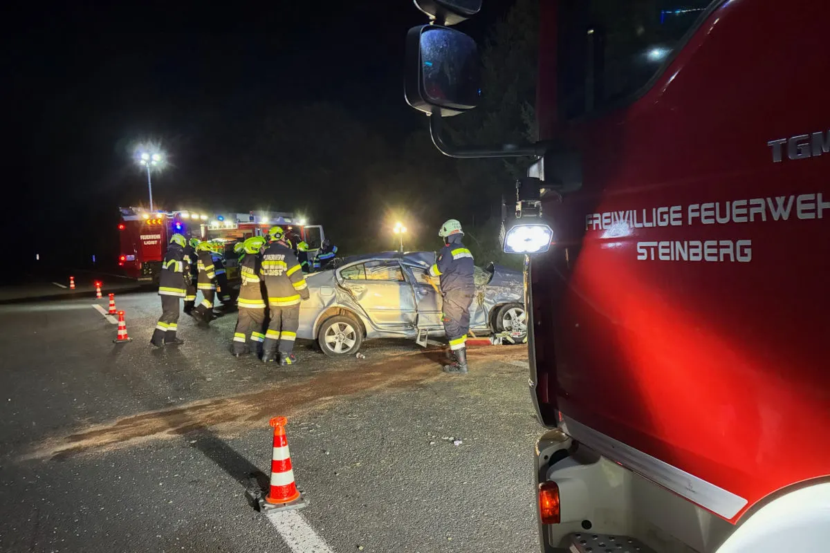 Eine Gruppe von Feuerwehrleuten steht in der Nacht um ein beschädigtes Auto auf einer Straße. Ein Feuerwehrfahrzeug ist sichtbar.