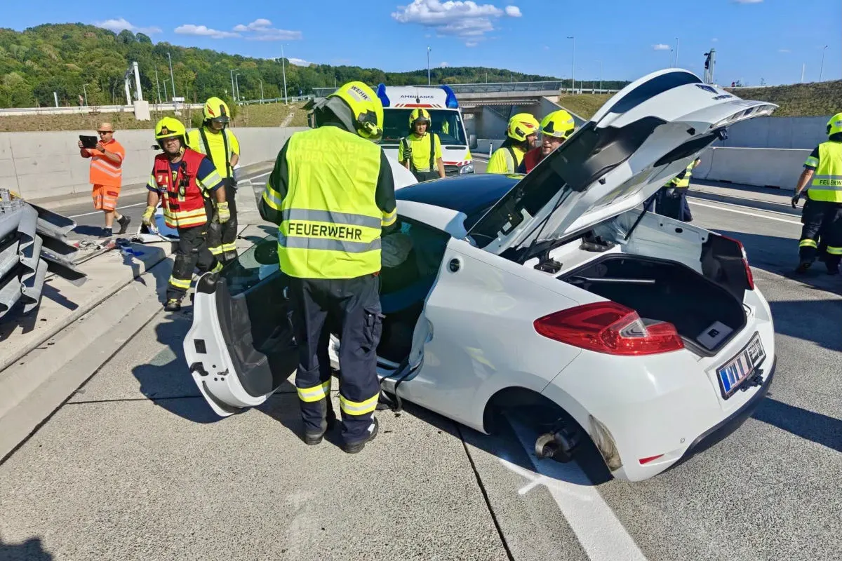 Mehrere Feuerwehrleute in gelben Helmen und Jacken arbeiten an einem weißen Auto, das auf der Autobahn steht. Im Hintergrund ist ein Rettungswagen zu sehen.
