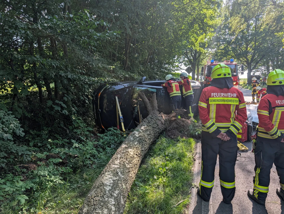 Ein Auto liegt zur Seite geneigt, nachdem es von einem umgestürzten Baum getroffen wurde. Feuerwehrleute arbeiten an der Bergung der Unfallstelle.